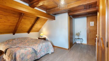 Ein Schlafzimmer mit schrägen Wänden, einem Bett mit Blumenmuster auf der Bettdecke, einem Nachttisch mit Lampe und einer Tür zu einem weiteren Raum.