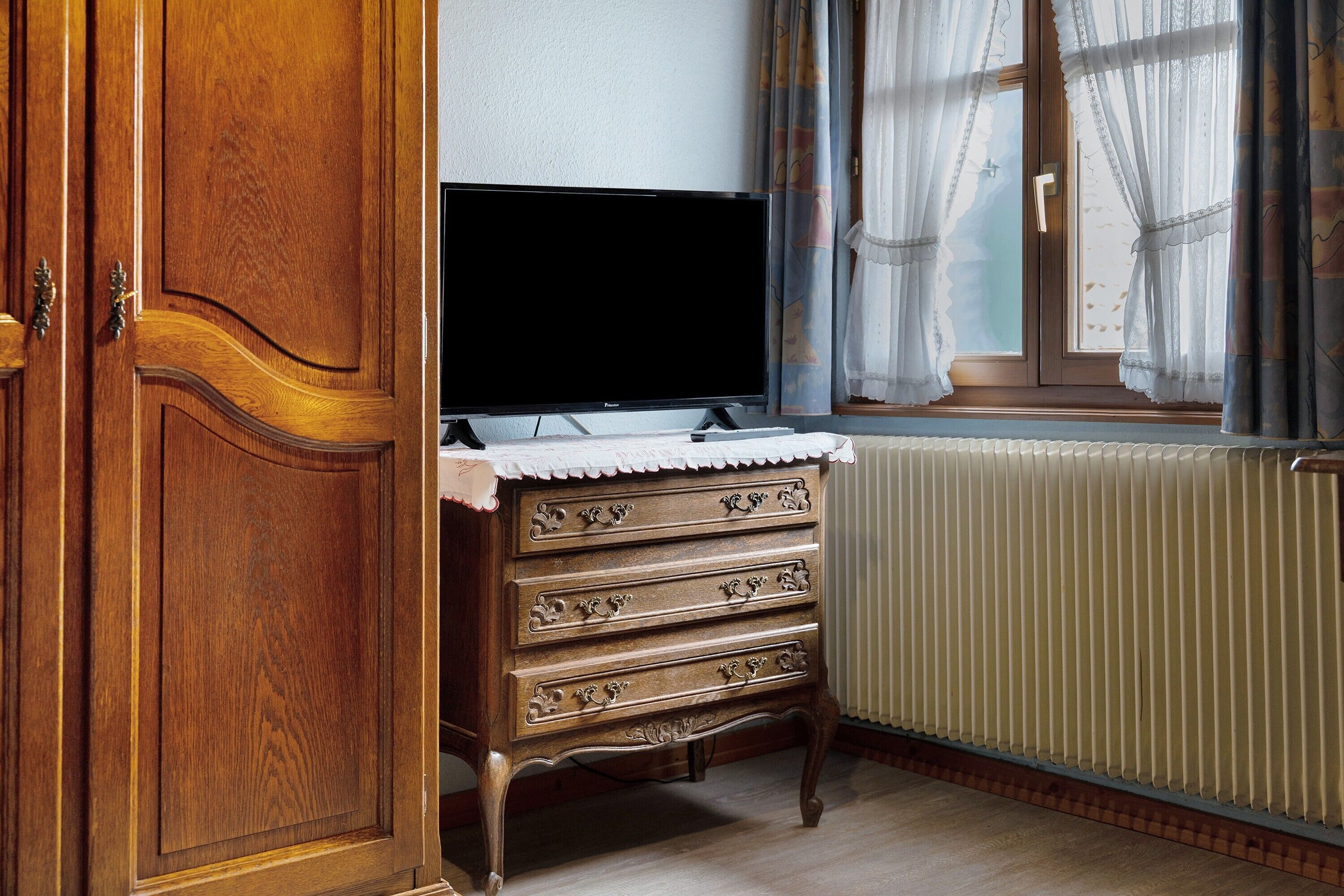 Ein Zimmer mit einem Holzschrank, einem Fernseher auf einer Kommode, einem Fenster mit Vorhängen und einem Heizkörper.