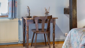 Um quarto com uma mesa de madeira e uma cadeira, um radiador, uma janela com cortinas e uma cama com colcha estampada.