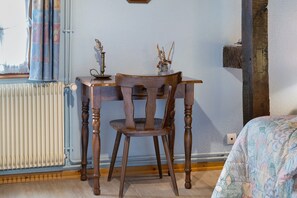 A room with a wooden table and chair, a radiator, a window with curtains, and a bed with a patterned bedspread.