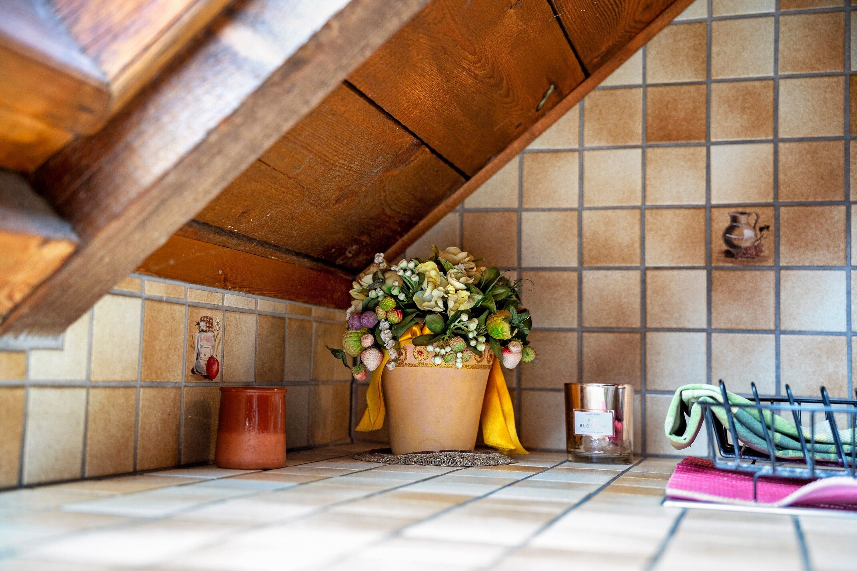 Un coin cuisine avec un mur recouvert de carreaux, un plafond en bois, une plante en pot, un petit vase et une étagère en métal où sont posés des torchons.