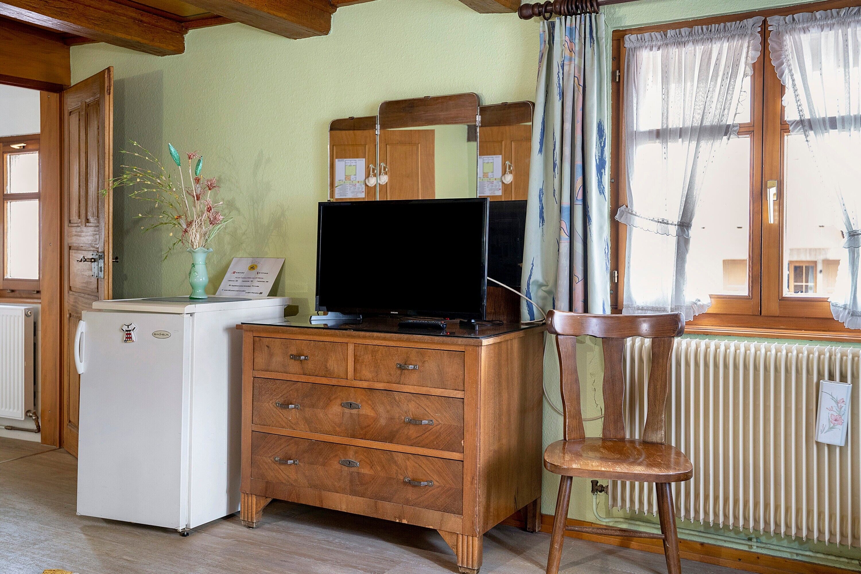 Een kamer met een houten dressoir, een televisie erboven, een kleine koelkast, een stoel en een raam met gordijnen.