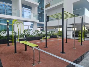 An indoor gym with exercise equipment, a red rubber floor, and a modern building with large windows in the background. - Vogue, Al Furjan Pearlz by Danube near Ibn Battuta Mall (Al Furjan)