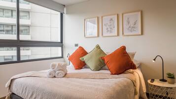 Habitación de hotel con un ventanal grande, una cama con una toalla enrollada y tres láminas botánicas enmarcadas en la pared.