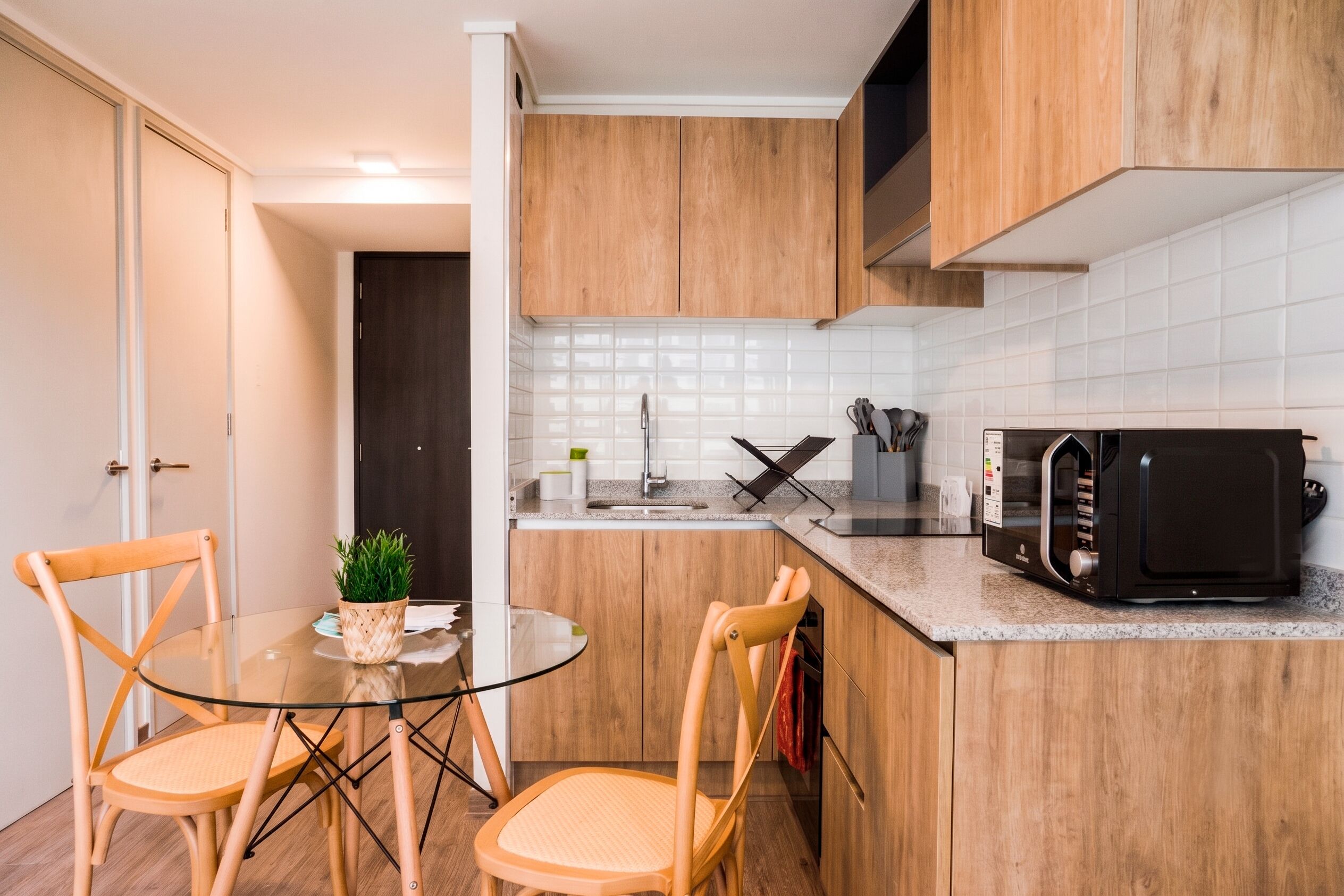 Cocina compacta con armarios de madera, microondas, lavamanos y una mesa de vidrio con sillas.