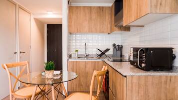 Cocina compacta con armarios de madera, microondas, lavamanos y una mesa de vidrio con sillas.