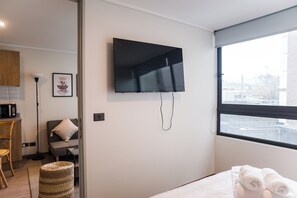 A modern hotel room with a flat-screen TV mounted on the wall, a bed with white bedding, a window with a view of the city, and a small kitchenette area. - Chic Las Condes 1BR1BA Pool & Gym (Las Condes)
