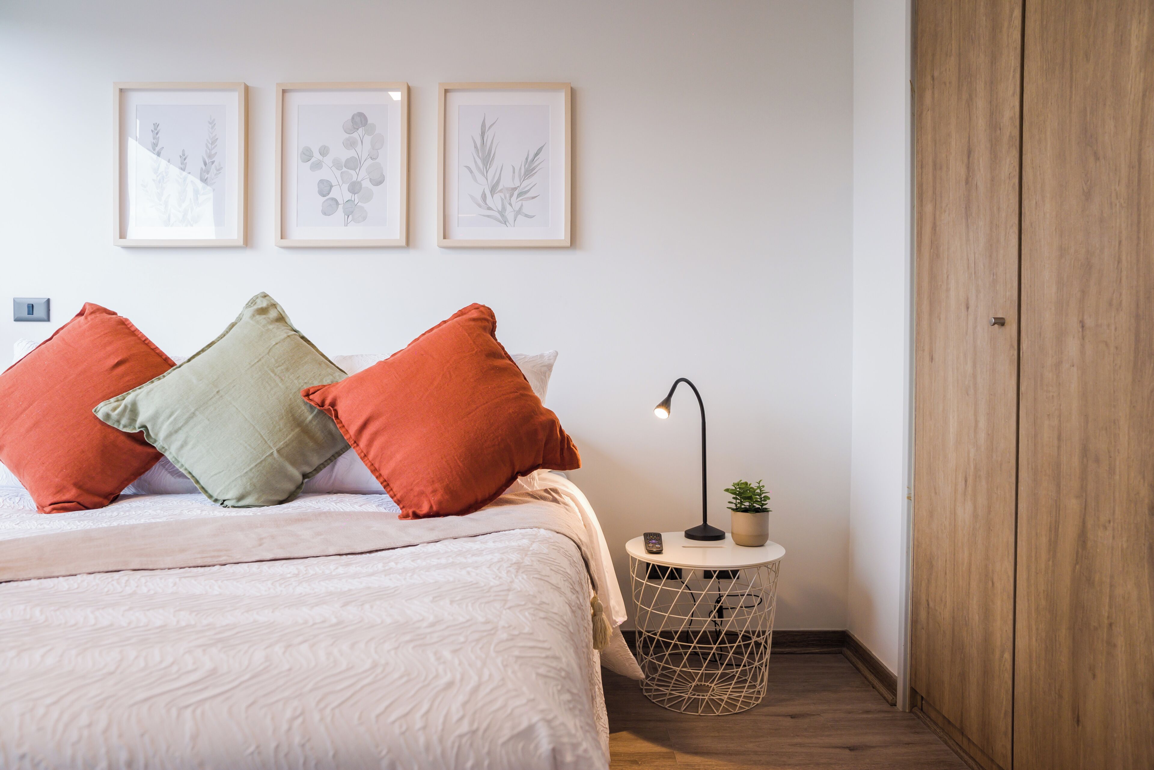 A bedroom with a bed, pillows, a bedside table with a lamp, a potted plant, and a wooden wardrobe.