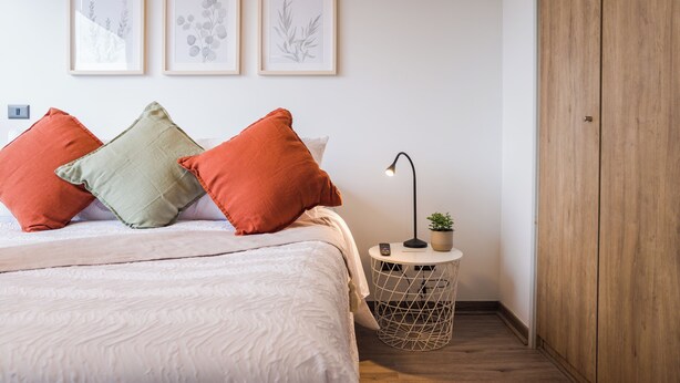 Een slaapkamer met een bed, kussens, een nachtkastje met een lamp, een kamerplant en een houten garderobe.