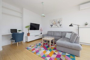 Un salon moderne avec un canapé gris, un téléviseur, un bureau avec un ordinateur, un tapis coloré et une plante en pot.