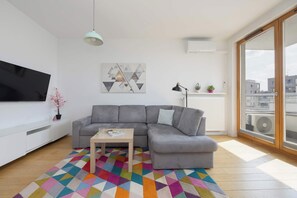 A modern living room with a grey sofa, a coffee table, a colorful rug, a flat-screen TV, and a large window with a city view. - Klimatisiertes Apartment | Parkplatz | Warschau-Wola (Warszawa)