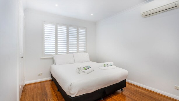Une chambre à coucher avec un lit, des murs blancs, une fenêtre avec des stores et un appareil de climatisation.