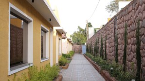 A pathway leading to a row of yellow buildings with windows and doors, flanked by a stone wall and greenery. - Sai Dhara Resort (Kasrawad)