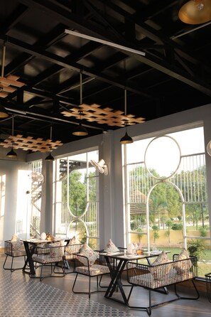 An indoor dining area with tables and chairs, large windows, and a view of greenery outside. - Sai Dhara Resort (Kasrawad)