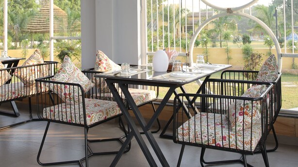 A dining area with a table set for four, surrounded by chairs with floral upholstery, large windows with circular frames, and a hanging light fixture. - Sai Dhara Resort (Kasrawad)