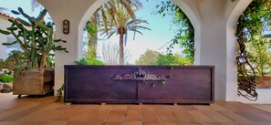A wooden bench with a decorative metal gate in a courtyard with arches, surrounded by potted plants and a palm tree.
