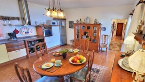 Een eetgedeelte met een houten tafel, stoelen en een schaal fruit. Er is een keuken met apparatuur en een kast met servies.