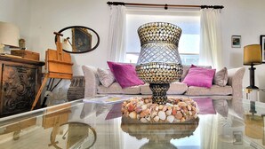 Een woonkamer met een glazen salontafel, een decoratieve lamp, een bank met kussens, een houten kast en een spiegel.