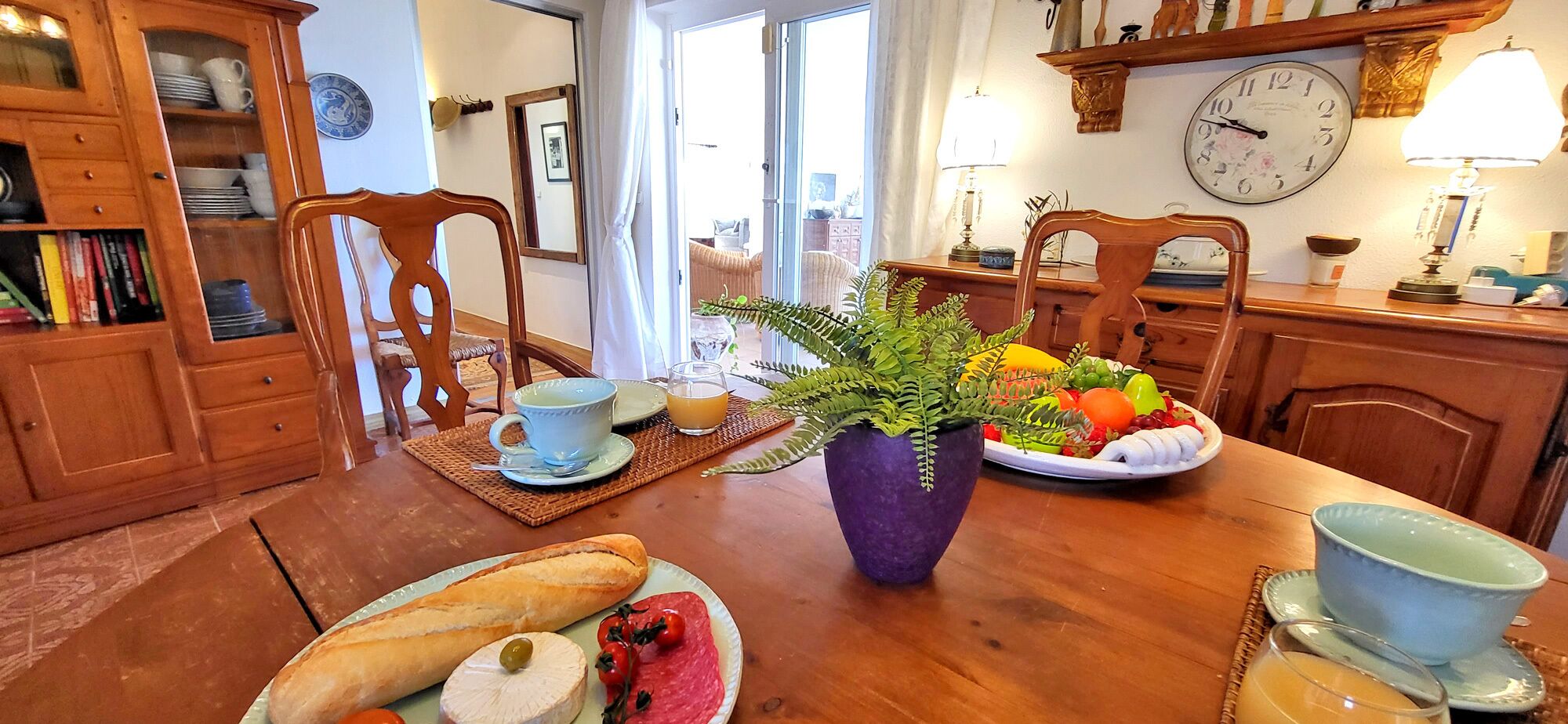 A dining area with a wooden table, chairs, a potted plant, a fruit bowl, a plate with a baguette, a cup, and a glass of orange juice.