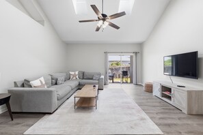 Un salon moderne avec un canapé d’angle, une table basse, un téléviseur fixé au mur et un ventilateur de plafond.