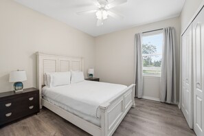 Une chambre à coucher comprenant un lit, des tables de chevet, une commode, un ventilateur de plafond et une fenêtre avec des rideaux.