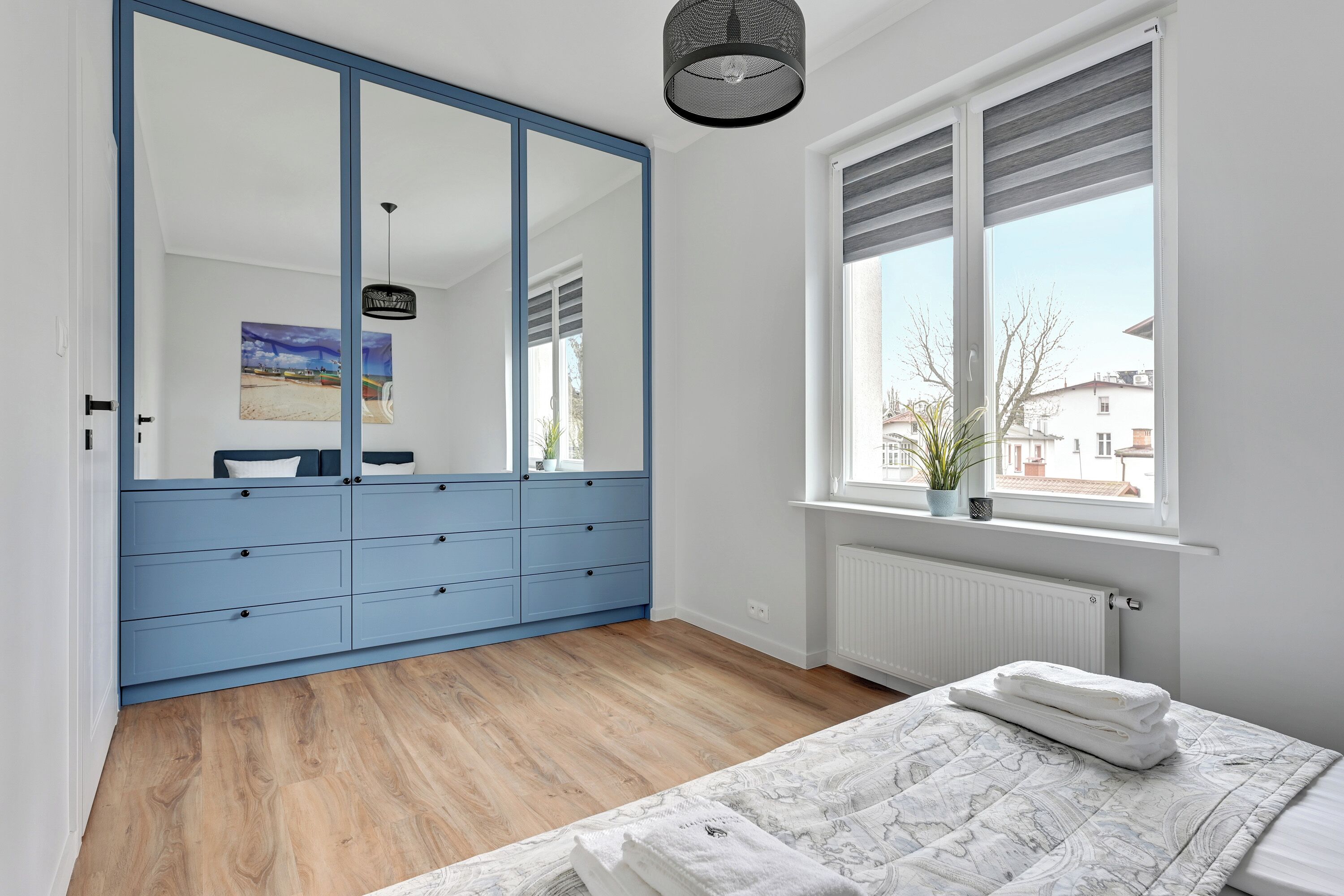 A bedroom with a large wardrobe, a bed with a patterned duvet, a window with blinds, and a hanging light fixture.