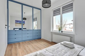 A bedroom with a large wardrobe, a bed with a patterned duvet, a window with blinds, and a hanging light fixture. - Ibiza - Sopot City Vibe Lion Apartments (Sopot)
