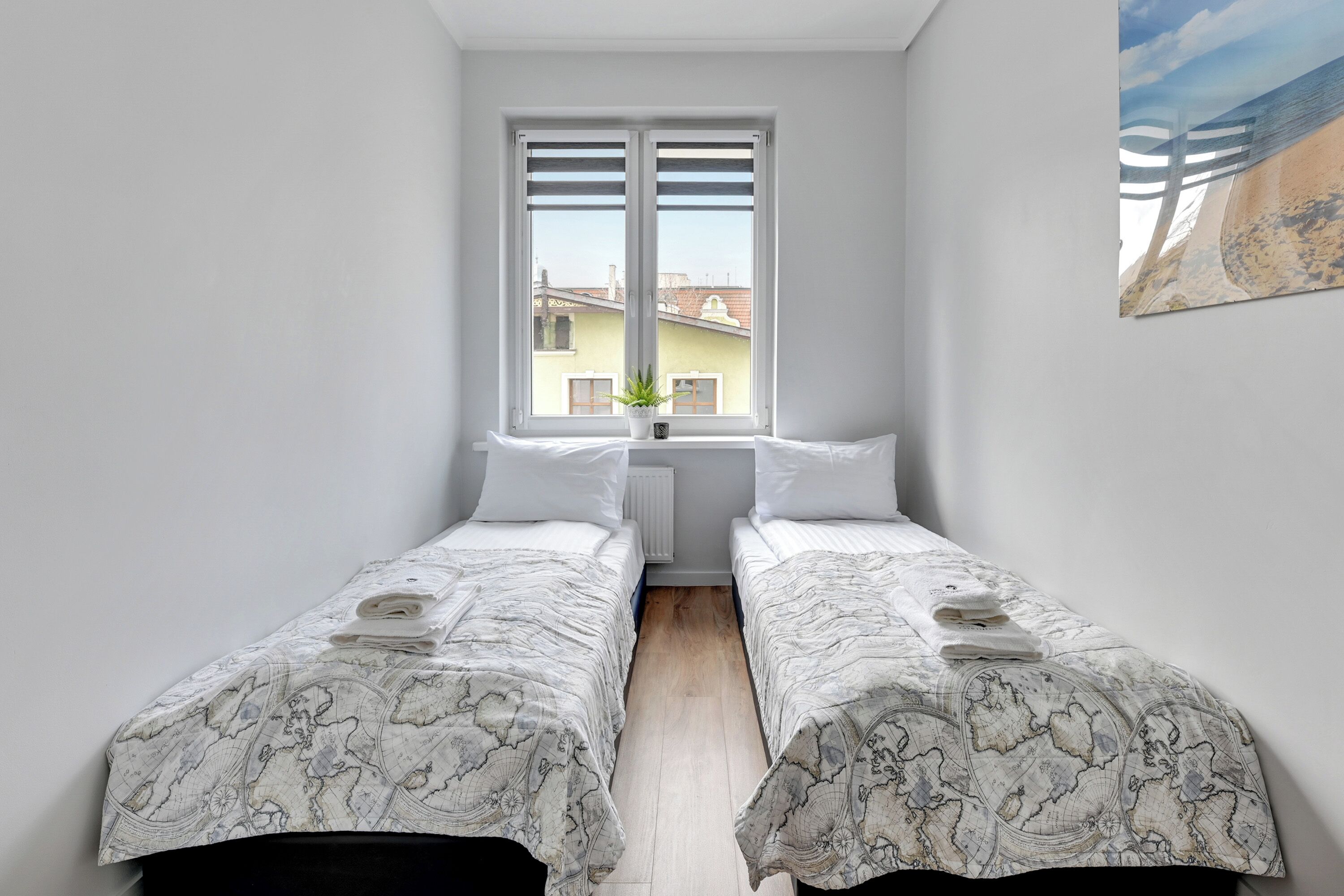 Una habitación pequeña y limpia con dos camas individuales, una ventana con persianas, un radiador y vistas a edificios.