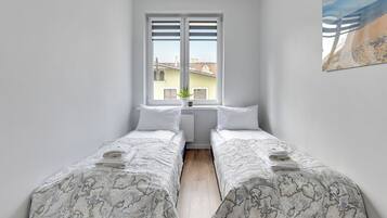 Una habitación pequeña y limpia con dos camas individuales, una ventana con persianas, un radiador y vistas a edificios.