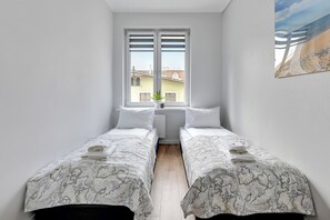 A small, clean room with two single beds, a window with blinds, a radiator, and a view of buildings outside. - Ibiza - Sopot City Vibe Lion Apartments (Sopot)