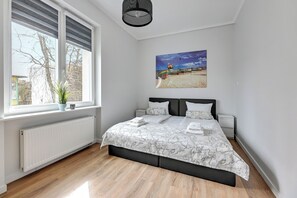 Um quarto com uma cama grande, uma janela com persianas, um radiador, um quadro de uma praia e um vaso de planta no parapeito.