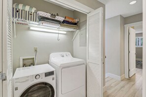 A laundry room with a front-loading washer and dryer, a shelf with folded towels, and a door leading to another room. - Sunset Rentals | Greens 170 Dog Friendly! (Hilton Head Island)