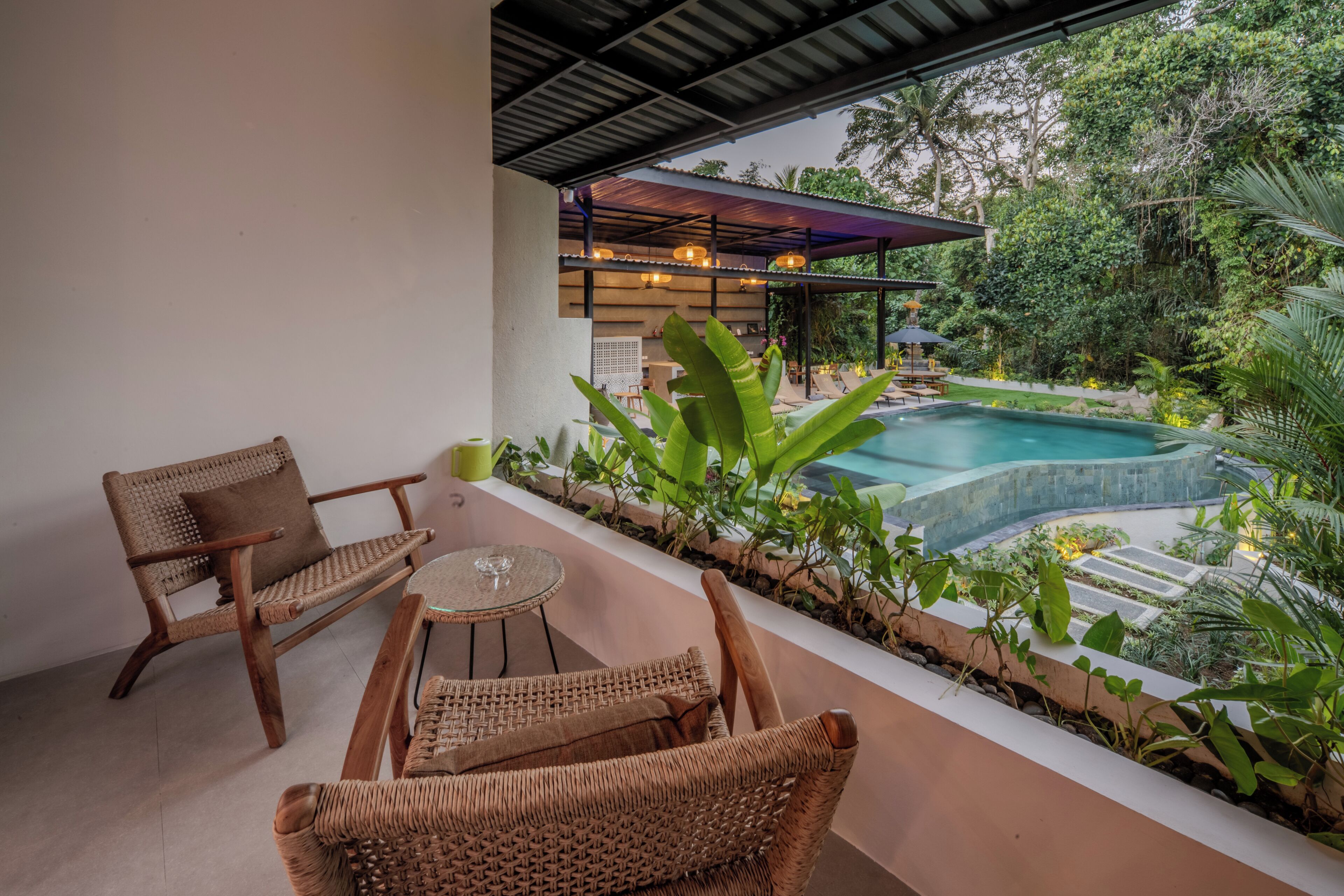 A patio with wicker furniture, a small table, and a pool surrounded by lush greenery.