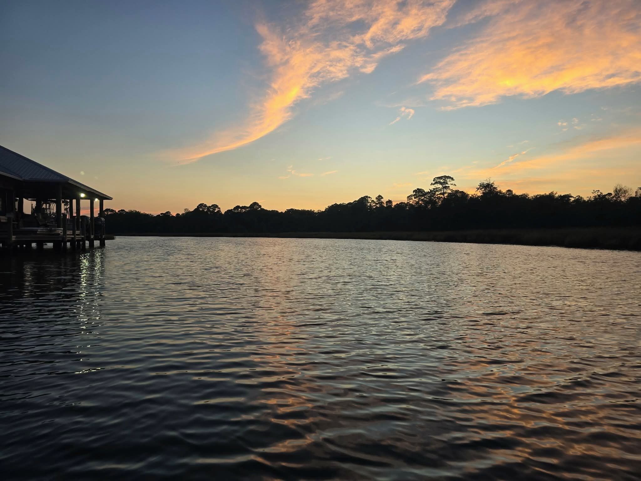 Propiedad frente al agua con un muelle y un edificio, rodeada de árboles y un cielo al atardecer.