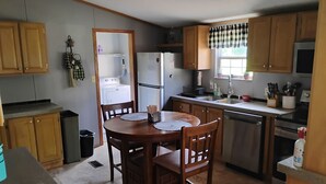 Une cuisine avec des meubles de rangement en bois, une table à manger avec des chaises, un réfrigérateur, un lave-linge et une fenêtre agrémentée d’une lambrequin à carreaux.