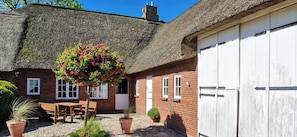 Una casa con tetto di paglia, facciata in mattoni, un portone garage bianco e un piccolo patio con tavolo e sedie.