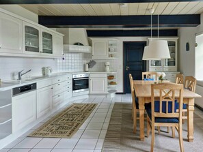 A modern kitchen with white cabinetry, a built-in oven, and a dining area with wooden chairs and a table. - Thatched cottage near the North Sea (Dagebüll)