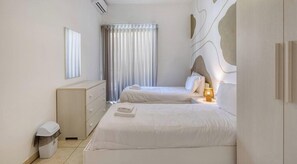 A hotel room with two beds, a bedside table with a lamp, a window with curtains, a waste bin, and a wall-mounted air conditioner. - Spinola Bay 3BR w/ beautiful Sunny Balcony & AC (San Ġiljan)