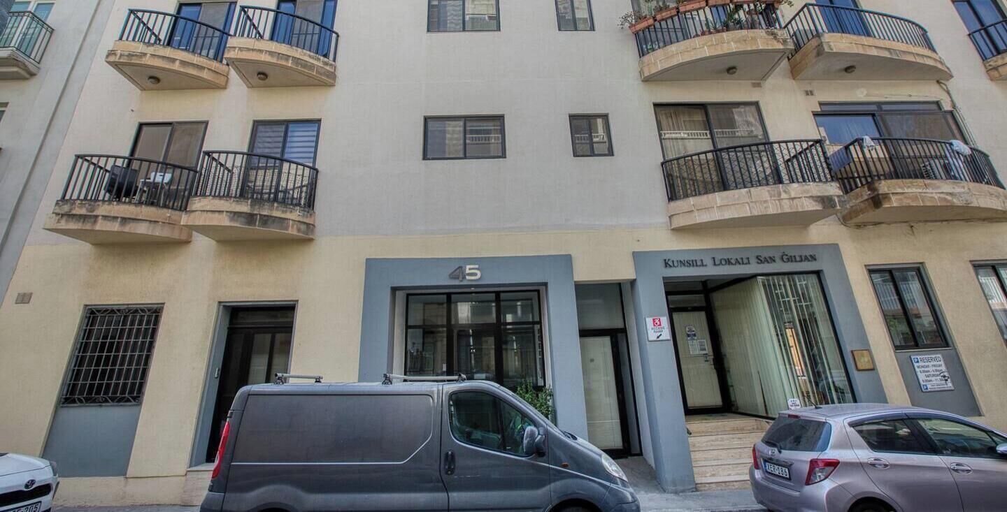 A multi-story building with balconies, a ground-level entrance, and a sign indicating the name 'Kunsill Lokali San Gilian.'