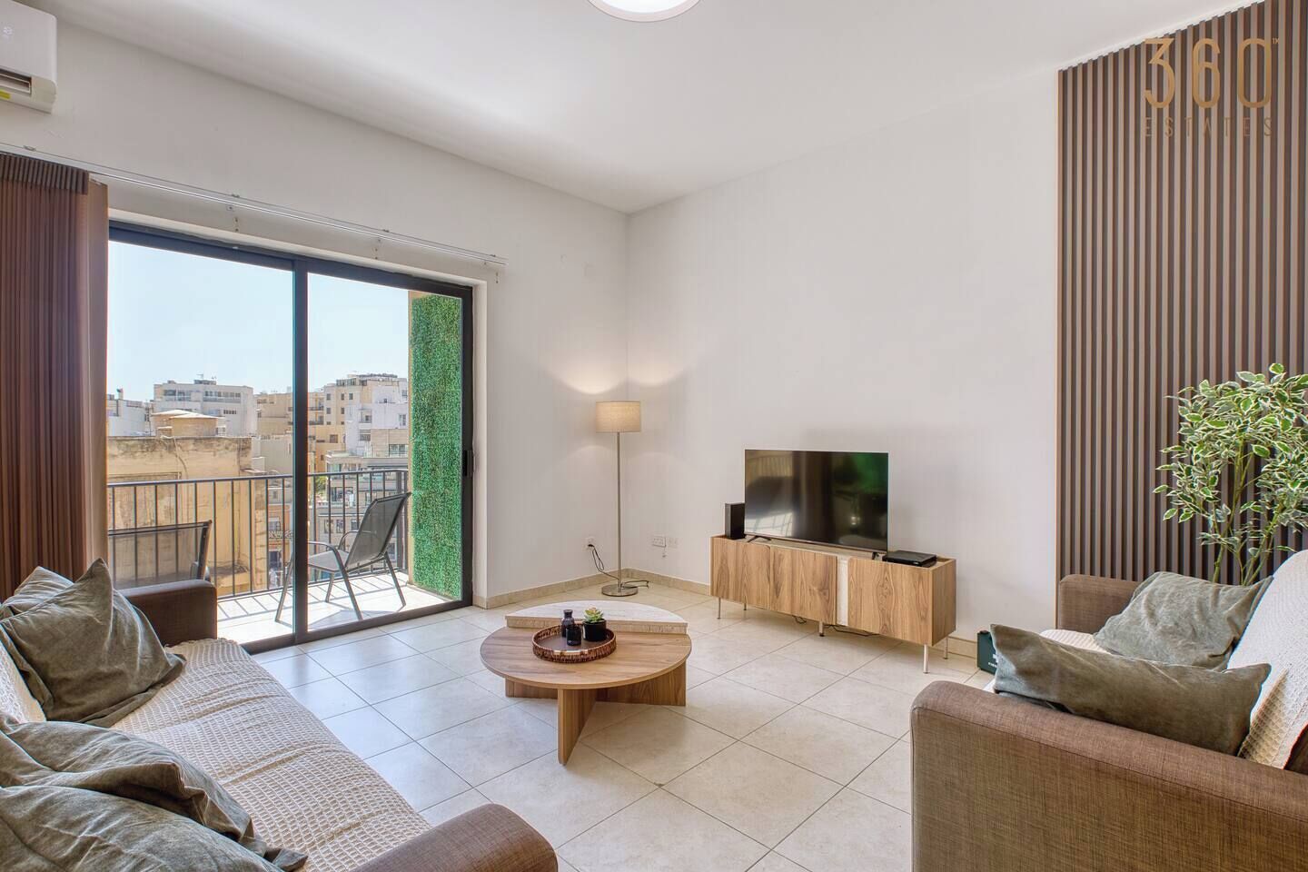 A modern living room with a sofa set, a coffee table, a TV on a wooden cabinet, and a large sliding glass door leading to a balcony.