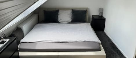 Un dormitorio pequeño y ordenado, con cama, mesita de noche, lámpara y radiador.