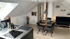 A modern kitchen with a central island, dining area with a wooden table and chairs, a flat-screen TV, and a ceiling with skylights.