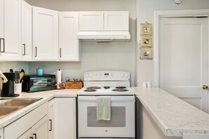A kitchen with white cabinets, a stove, a microwave, and a sink. - Sleep 4 in this bright Pensacola cottage just minutes from Perdido Key's white sands and the National Naval Aviation Museum. Full kitchen, in-unit laundry, comfy queen bed, and a picnic-table yard. Your easy Gulf Coast home base awaits! (Pensacola)