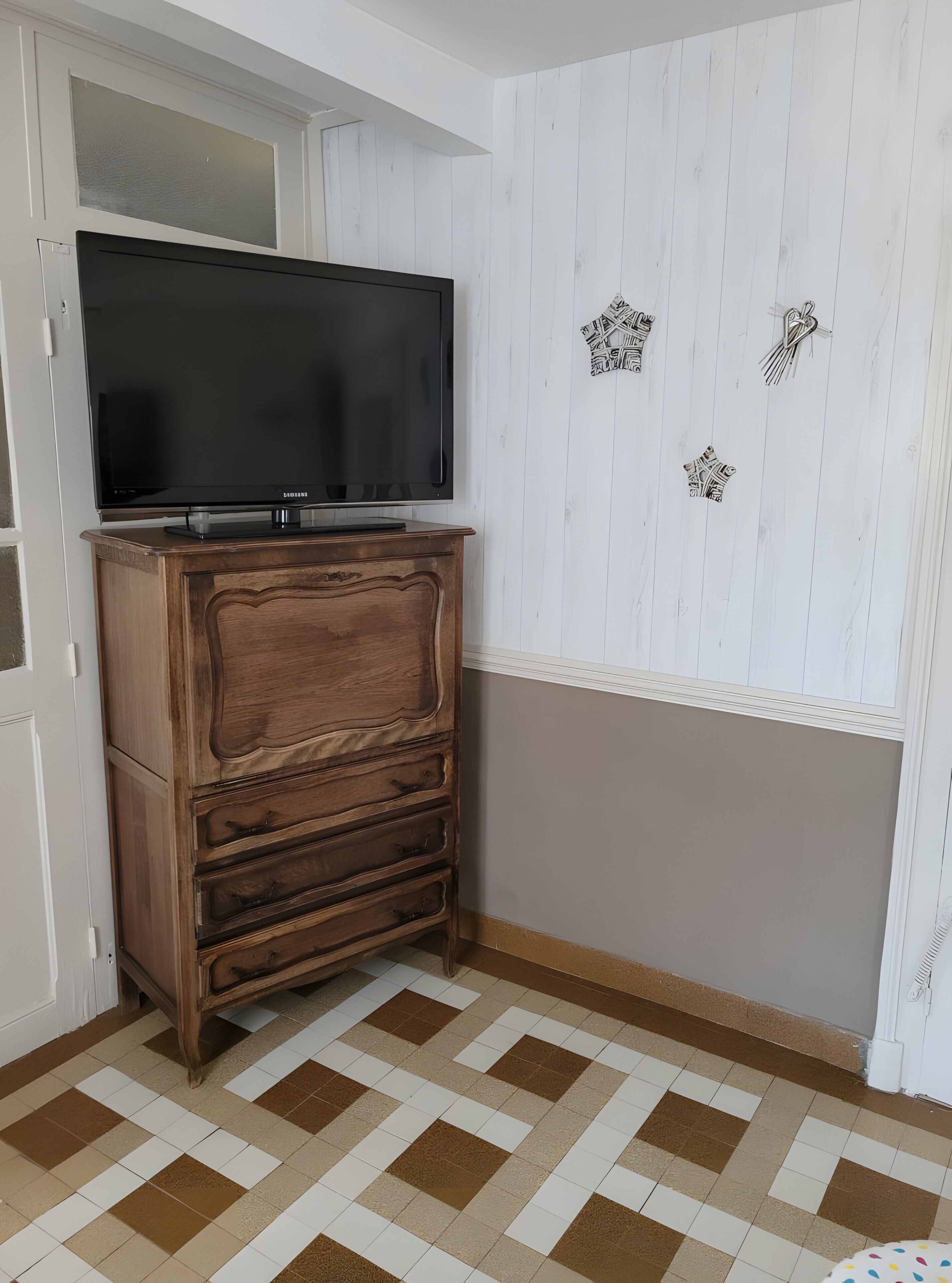 Une pièce avec un téléviseur posé sur un meuble en bois, un sol à motifs et un mur blanc agrémenté d’éléments décoratifs.