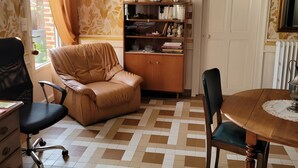 Ein Zimmer mit Schachbrettboden, einem Ledersofa, einer Holzkassette mit Büchern, einem Esstisch mit Stühlen und einer Wand mit Muster.