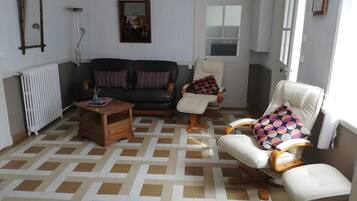Uma sala de estar com sofá, duas poltronas, uma mesa de centro, um radiador e uma janela com cortinas.
