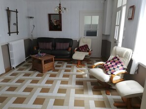 A living room with a sofa, two armchairs, a coffee table, a radiator, and a window with curtains.