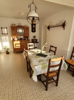 A dining area with a table set for four, surrounded by wooden chairs. A chandelier hangs above the table, and a wooden cabinet with a clock is in the background.