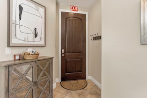 A hallway with a wooden door, a framed abstract painting, a basket on a console table, and a doormat. - Cactus Rose at Coral Ridge | Sage Vacations (Washington)