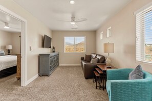 A room with a bed, a sofa, a small table, a cabinet, a ceiling fan, a window with blinds, and a view of a desert landscape. - Cactus Rose at Coral Ridge | Sage Vacations (Washington)